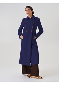 Cappotto doppiopetto blu navy con bottoni dorati, dotato di un colletto a risvolto e tasche laterali, abbinato a pantaloni larghi marroni e scarpe metalliche.