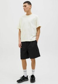 Tricou din bumbac de culoare galben deschis, cu mâneci scurte, asociat cu pantaloni scurți cargo negri care au un buzunar lateral. Modelul poartă adidași negri și șosete albe.