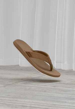 Infradito marrone in memory foam con cinturino in tessuto, fluttuante sopra un pavimento in legno chiaro con tende bianche trasparenti sullo sfondo.