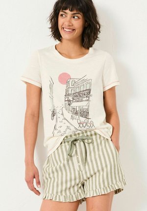 Cremfarbendes T-Shirt mit einem Grafikmotiv einer Straßenansicht, kombiniert mit grün-weiß gestreiften Shorts mit einem rüschierten Saum und Bindegürtel.