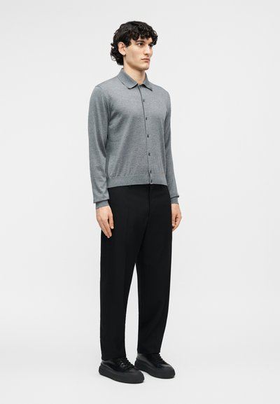 Jeune homme aux cheveux bouclés et foncés portant une chemise grise à manches longues boutonnée, un pantalon noir et des baskets noires, debout devant un fond uni.