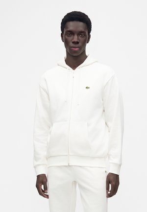 Joven con sudadera blanca con cremallera y pantalones blancos a juego con un pequeño logo verde en el pecho y los pantalones, de pie frente a un fondo liso.