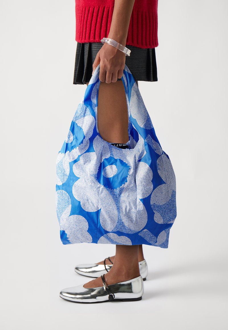 Reusable blue bag featuring large white floral patterns, durable material, and cutout handles for easy carrying. Silver shoes visible below.