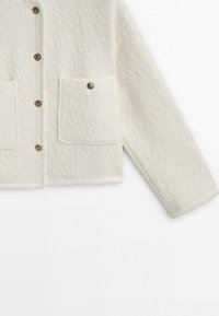 Veste en laine blanche texturée avec manches longues, fermeture à boutons sur le devant et deux poches carrées chacune ornée d'un bouton décoratif.