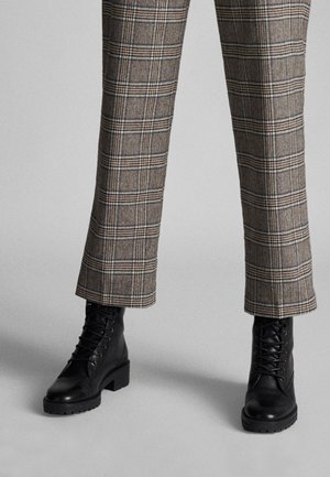 Jambes portant un pantalon droit à carreaux marron associé à des bottines noires à lacets sur fond gris.