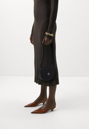 Borsa a spalla in pelle nera con una forma curva, caratterizzata da un logo argentato. Abbinata a un vestito marrone a coste e tacchi a punta marroni.