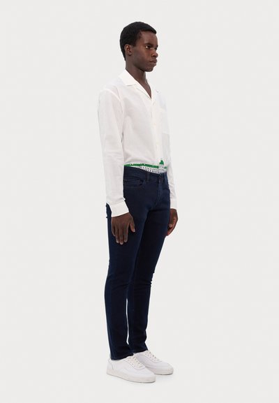 Weißes Hemd mit Knöpfen und einer Tasche, dunkelblaue Jeans und weiße Schuhe. Das Hemd hat einen grünen Akzent an der Taille. Glatte Stofftextur.