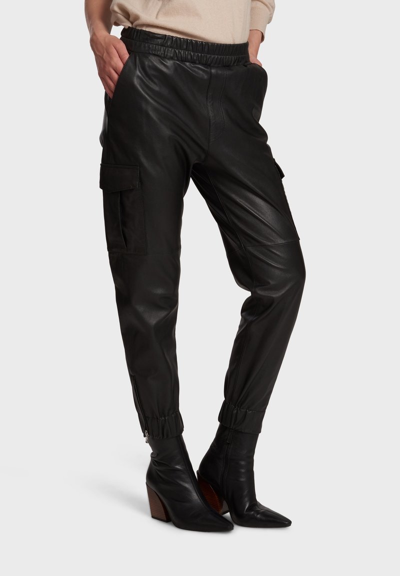 Svarta läder cargobyxor med elastisk midja, sidofickor och dragkedja vid bensluten. Modellen bär spetsiga svarta klackskor.