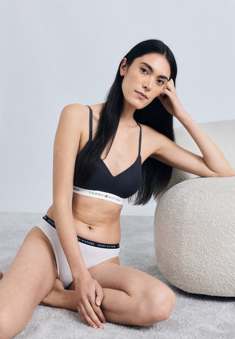 Czarny biustonosz typu bralette z cienkimi ramiączkami i paskiem z logo; jasnoszare majtki z wysokim stanem z kontrastowym paskiem z logo; gładka tkanina; swobodna poza.