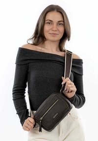 Hnědá texturovaná crossbody taška se zipovou kapsou, zvýrazněná vzorovaným popruhem a zlatým logem, kterou drží modelka oblečená v černém topu.