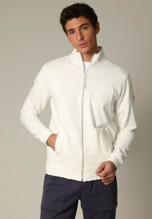 Veste blanche à fermeture zippée, avec un col haut, des manches longues et une texture subtile. Elle présente une poche avant et un détail de fermeture éclair multicolore.