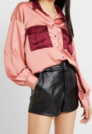 Vrouw draagt een roze satijnen blouse met knopen, donkerrode zakken en kraag, gecombineerd met hooggetailleerde zwarte leren shorts met riem.