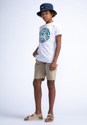 Witte T-shirt met kleurraf rond graphic, beige shorts, grijze sandalen met gesp, en blauwe bucket hat. Effen lichte achtergrond.