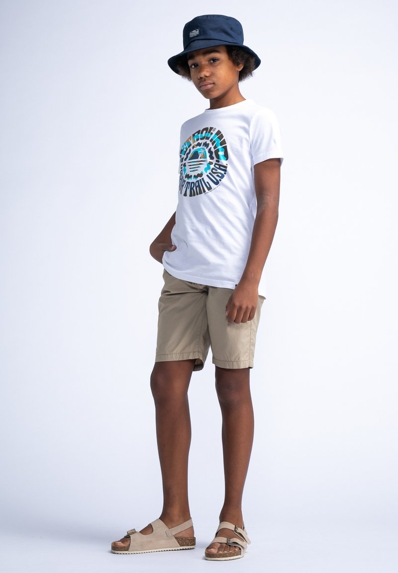 Witte T-shirt met kleurraf rond graphic, beige shorts, grijze sandalen met gesp, en blauwe bucket hat. Effen lichte achtergrond.