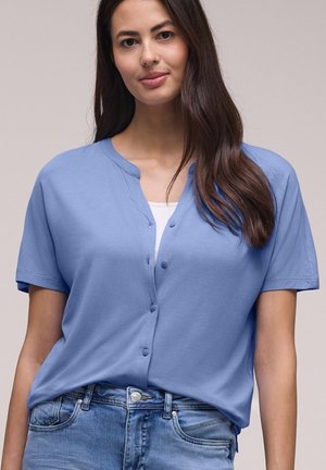 Ung kvinde med langt mørkt hår iført lyseblå kortærmet knappet skjorte over hvid top og blå jeans, stående mod en ensfarvet baggrund.