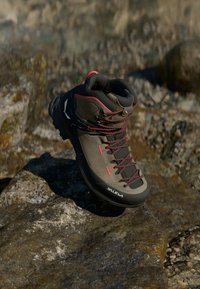 Šedá a černá turistická bota s červenými akcenty, texturovaný povrch a robustní podrážka, odpočívající na kamenitém povrchu. Obsahuje zesílené okopové otvory a tkaničky.