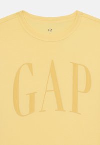 Žluté bavlněné tričko s velkým, mírně tmavším logem "GAP" moderním písmem. Kolo výstřih a kratké rukávy. Jednoduchý design.