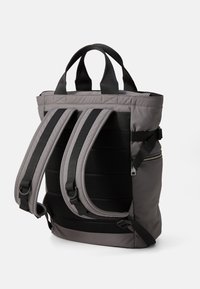 Sac à dos gris avec un extérieur matelassé, un dos en mesh noir et des sangles réglables. Il possède deux poignées supérieures et une poche latérale zippée.