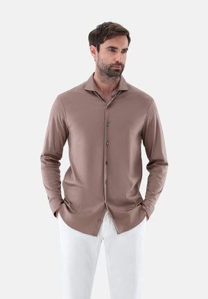 Camicia button-up a maniche lunghe in tessuto marrone tenue, con colletto a punta, texture liscia e bottoni scuri, abbinata a pantaloni bianchi.