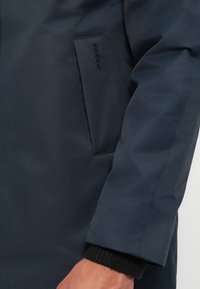 Sötét tengerészkék vízálló kabát sima felülettel, oldalzsebbel és bordás fekete mandzsettákkal. Barbour márkajelzés a zsebén.