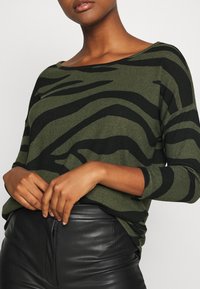 Olivgrüner Pullover mit schwarzen Zebrasstreifen, ausgestattet mit einem weiten Ausschnitt und langen Ärmeln, kombiniert mit einer schwarzen Lederhose.