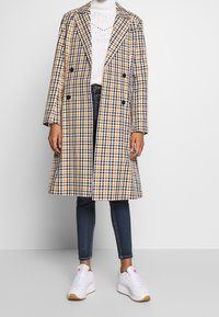 Rutigt mönstrad kappa i gult, marinblått och vitt; lång dubbelknäppt design med stora slag. Bärs över en vit polotröja och blå jeans, med vita sneakers.