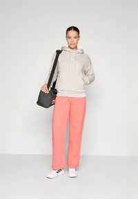 Ung kvinna i en beige huvtröja, korallfärgade sweatpants, vita sneakers, som bär en svart axelväska mot en ljus, enkel bakgrund.