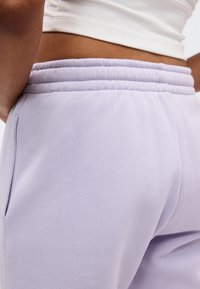 Κοντινό πλάνο ατόμου που φοράει ανοιχτόμωβ φόρμα με ελαστική μέση και λευκό κοντό τοπ, εμφανίζοντας τη μέση και την πλαϊνή τσέπη.