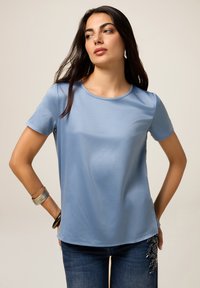 Top scurt de culoare albastru deschis, cu un guler rotund, textură netedă și o croială ușor lejeră. Asortat cu blugi cu accente decorative.