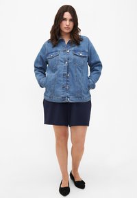 Heled sinine teksajakk, millel on nööbid, kaks rinnataskut ja pikad varrukad, kantud tumesiniste lühikeste pükste ja mustade lamekingade peale. Sile teksakanga tekstuur.