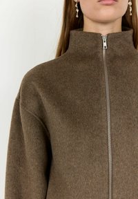 Hnedá fleecová bunda na zips s vysokým golierom, so strieborným zipsom a jemným, textúrovaným povrchom.