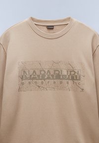Sweat-shirt beige à col rond avec un rectangle imprimé craquelé affichant « NAPAPIJRI » en grandes lettres et « Geographic » en plus petites lettres en dessous.