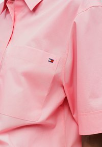 Nærbillede af lyserød kortærmet skjorte med brystlomme med en lille rektangulær flaglogo i rød, hvid og blå.