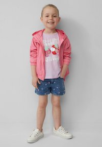 Rosa Zip-Jacke über einem helllila T-Shirt mit Obstgrafiken, kombiniert mit Denimshorts mit Blumenmustern und weißen Sneakers.
