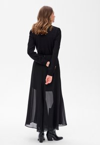 Zwarte lange rok met doorschijnende overlay, gecombineerd met een getailleerd zwart top met gestructureerde mouwen. Accessoires zijn onder andere een armband en enkelboots.