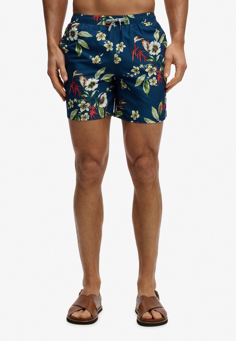 Personne aux jambes nues portant un short de bain bleu à motifs floraux et des sandales en cuir marron, debout devant un fond blanc uni.
