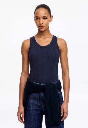 Jeune femme aux cheveux foncés attachés en arrière, portant un haut côtelé sans manches bleu marine et un jean foncé avec un pull noir noué autour de la taille.