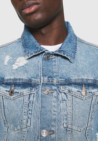 Světle modrá džínová bunda s opranými detaily, stříbrnými knoflíky, dvěma předními kapsami a špičatým límcem. Texturovaná látka s viditelným prošitím.