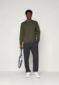 Felpa verde con collo a giro, abbinata a pantaloni grigi scuri e affusolati. Il modello tiene in mano una racchetta da tennis e indossa scarpe da ginnastica bianche.