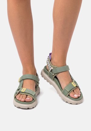 Piedi con sandali verdi da esterno con cinturini regolabili e suole spesse beige, in piedi su uno sfondo bianco.