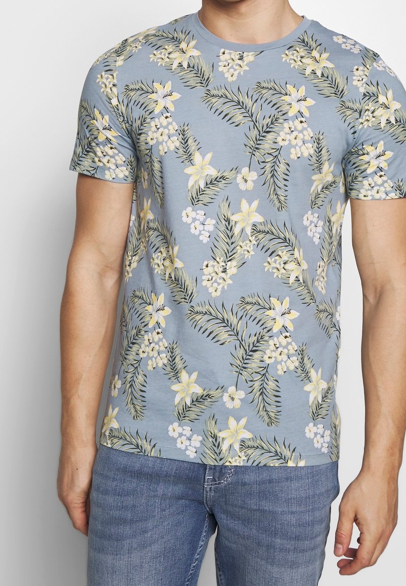 Světlemodré tričko s krátkým rukávem s květinovým vzorem, na kterém jsou žluté květy a zelené listy. Vyrobeno z měkké bavlněné látky.