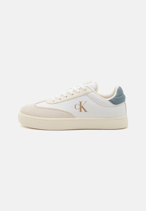 Zapatilla de lona blanca con acentos de ante beige, detalle de talón azul claro, punta redonda, cordones blancos y logo dorado en el lateral.