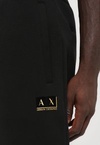 Tuvuma tuvums, uz kura atrodas roka blakus melniem Armani Exchange biksēm ar zelta "A|X Armani Exchange" logotipu netālu no kabatas.