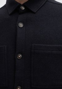 Gros plan d'une veste en laine bleu marine foncé avec un col, une fermeture boutonnée devant et deux grandes poches poitrine.