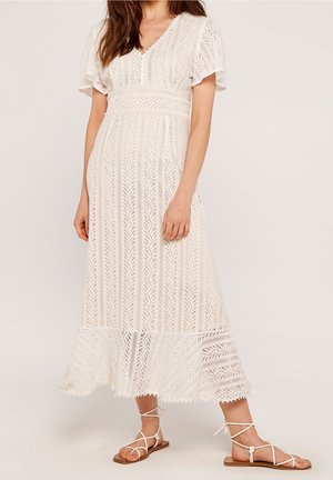 Mujer con vestido midi blanco de encaje con mangas tipo volante y sandalias planas con tiras sobre un fondo neutro.