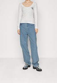 Långärmad vit topp med djup V-ringning och liten svart logotyp; matchas med ljusblå vida jeans och blanka svarta klackskor.