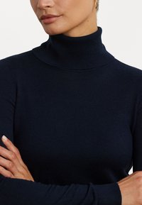 Tummansininen poolopaita, joka on valmistettu pehmeästä neulemateriaalista. Ominaisuudet ovat tiukka muotoilu ja pitkät hihat, joissa on ribbikuviointi kauluksessa ja hihoissa.
