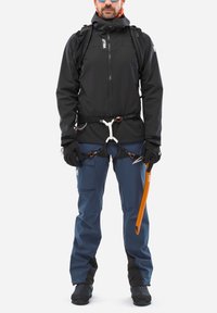 Man wearing black jacket, blue climbing pants, harness, gloves, sunglasses, and carrying an orange ice axe and backpack.