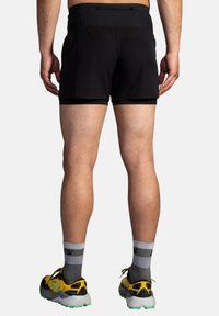 Zwarte sportshorts met een gladde textuur en een aansluitend ontwerp; gedragen met gele en grijze hardloopschoenen met een gestructureerde zool.