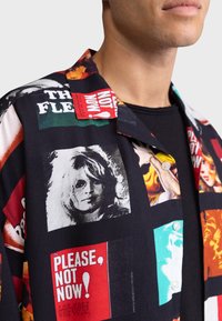 Zbliżenie czarnej koszuli z guzikami, ozdobionej kolorowymi grafikami, w tym napisem "Proszę, nie teraz!" oraz czarno-białym portretem kobiety.